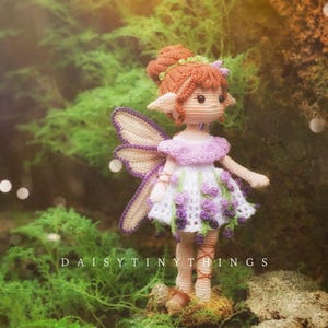 Könnte beinhalten: Eine handgefertigte Häkelfee-Puppe mit einem weißen und lila Kleid, lila Flügeln und rotem Haar. Die Puppe steht auf einer moosigen Oberfläche. Der Text "DAISY TINY THINGS" ist sichtbar.