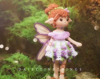 May include: A handmade crochet fairy doll with a white and purple dress, purple wings, and red hair. The doll is standing on a mossy surface. The text "DAISY TINY THINGS" is visible.