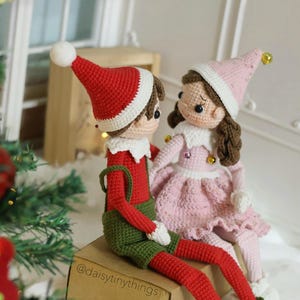 May include: A crocheted elf couple wearing red and green clothing and pink and white clothing. The elf couple is sitting on a brown box with the text "@daisytinythings" on it.