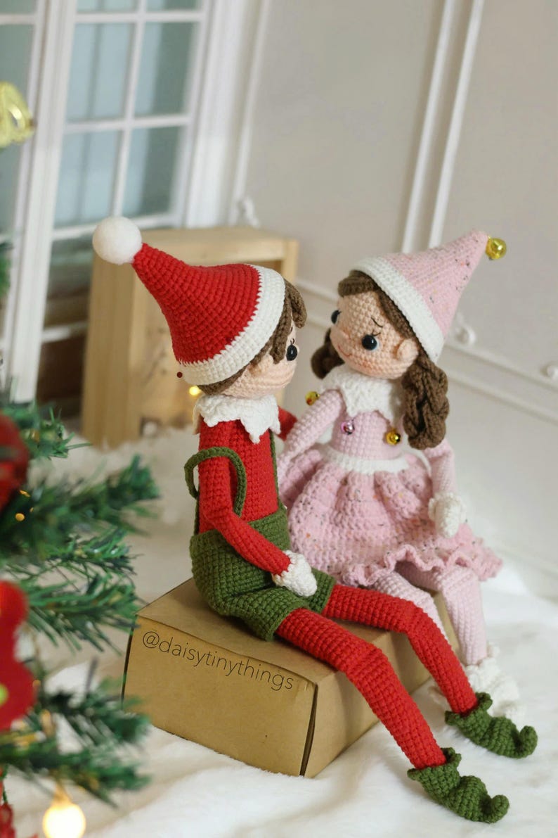May include: A crocheted elf couple wearing red and green clothing and pink and white clothing. The elf couple is sitting on a brown box with the text "@daisytinythings" on it.