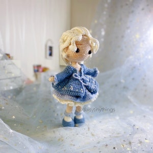 May include: A crocheted doll with blonde hair, wearing a blue cardigan and a white dress with blue accents. The doll is standing on a blue sparkly surface.