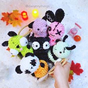 May include: A collection of seven crocheted plush toys in a basket. The toys are various colors, including green, pink, black, and white. The toys have stitched facial features and some have small details like buttons or yarn accents. The toys are arranged in a basket with a brown handle. The basket is sitting on a white surface. The text "@daisytinythings" is visible in the image.