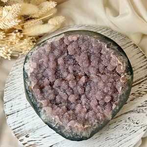 May include: A geode with a dark grey exterior and a cluster of sparkling, light purple crystals inside. The geode is displayed on a white, distressed wooden tray, with dried flowers in the background.