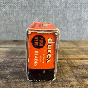 May include: A vintage Durex double edge blades box. The box is orange and black with the Durex logo and text that reads "Push Here Blades". The box is rectangular and appears to be made of cardboard.
