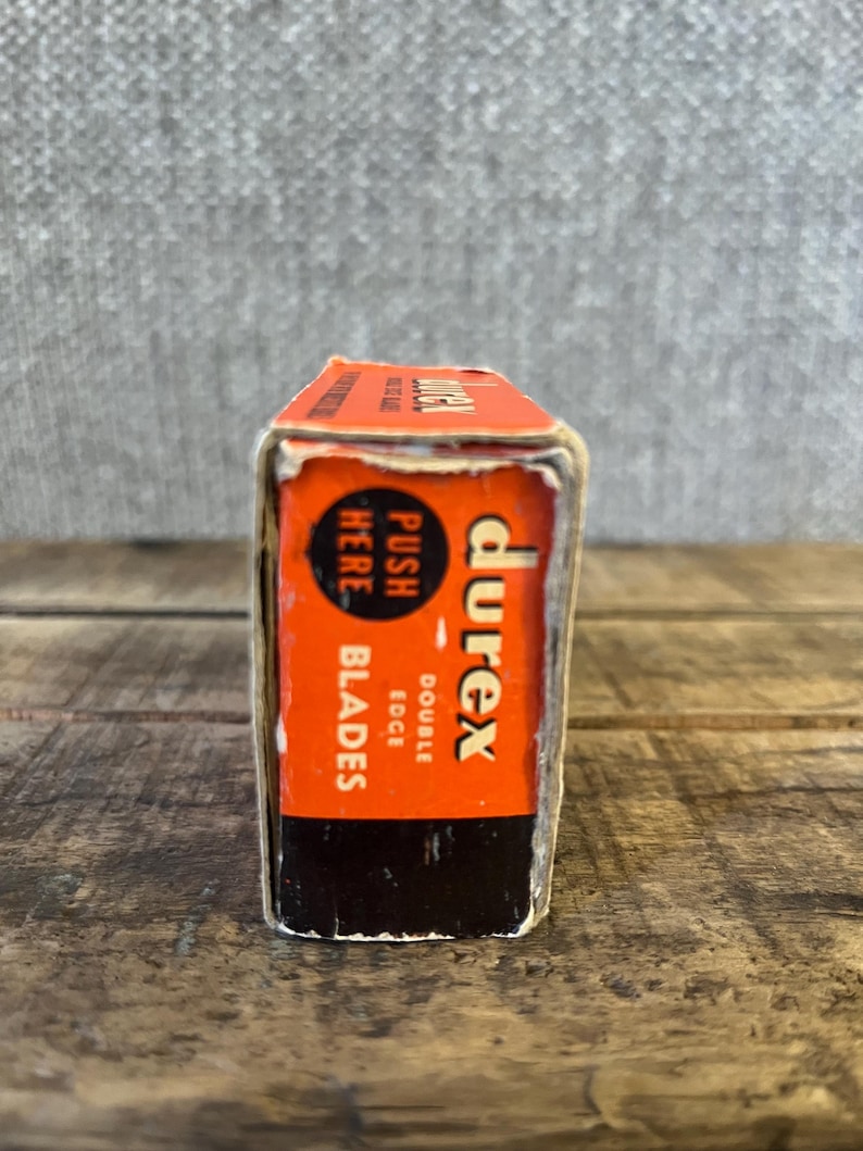 May include: A vintage Durex double edge blades box. The box is orange and black with the Durex logo and text that reads "Push Here Blades". The box is rectangular and appears to be made of cardboard.