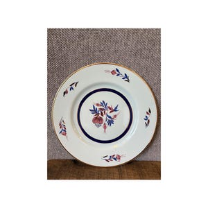 May include: White ceramic plate with a gold rim and a central floral design in red, blue, and pink. The plate features a dark blue ring around the floral arrangement and smaller floral accents around the edge.