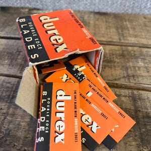 May include: An open cardboard box of Durex double edge blades. The box is orange and black with the brand name in white. Several individual blade packages are visible, also orange and black.