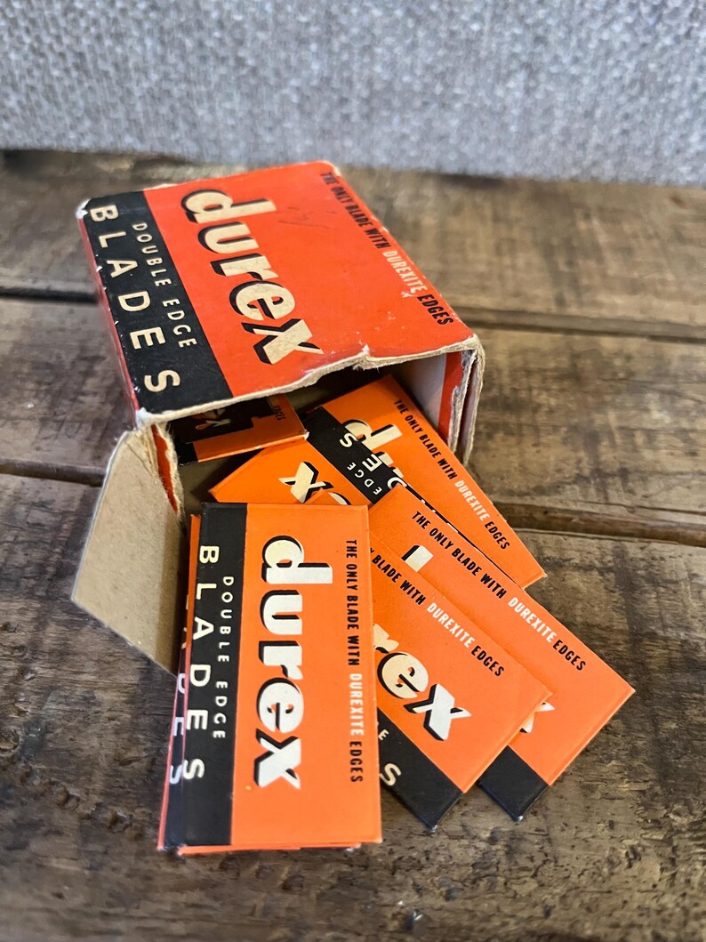 May include: An open cardboard box of Durex double edge blades. The box is orange and black with the brand name in white. Several individual blade packages are visible, also orange and black.