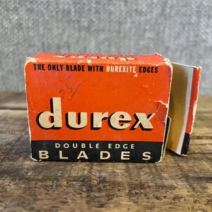 May include: Vintage Durex double edge blades box. The box is orange with black and white text. The text on the box reads "THE ONLY BLADE WITH DUREXITE EDGES" and "DOUBLE EDGE BLADES". The box is on a wooden surface.