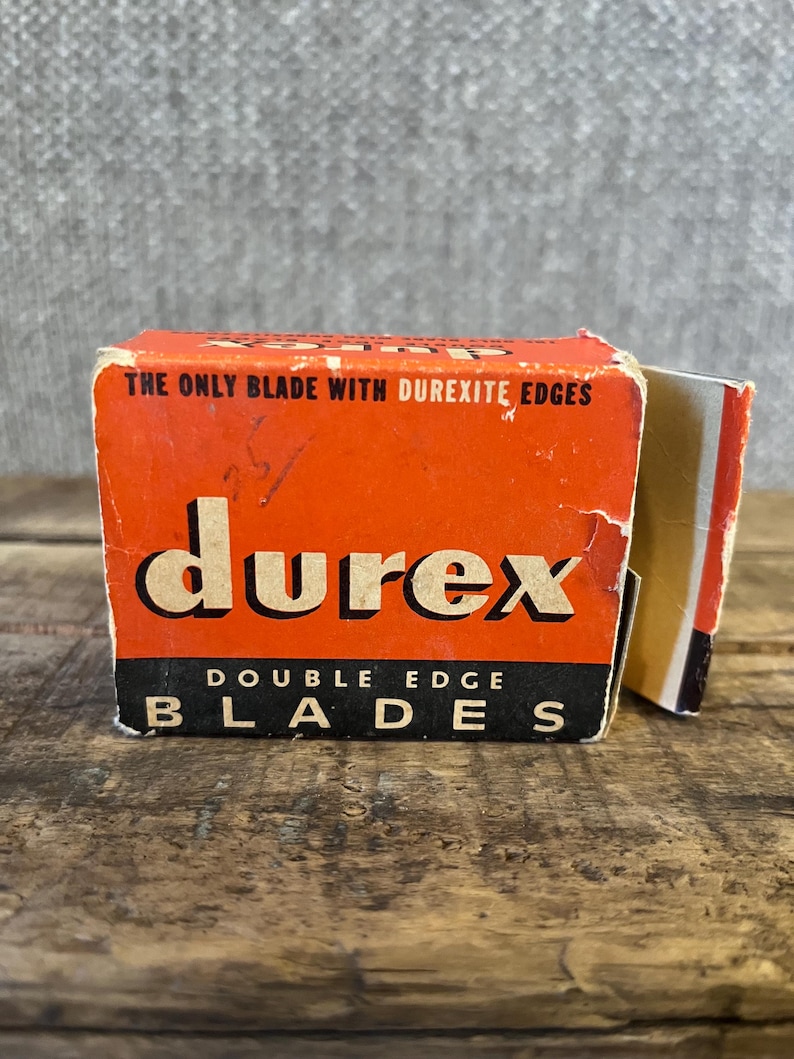 May include: Vintage Durex double edge blades box. The box is orange with black and white text. The text on the box reads "THE ONLY BLADE WITH DUREXITE EDGES" and "DOUBLE EDGE BLADES". The box is on a wooden surface.