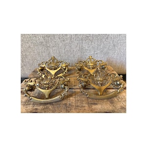 May include: Two antique brass drawer pulls with ornate floral and scrollwork designs. The pulls have a curved handle and decorative top piece. They are displayed on a weathered wooden surface.