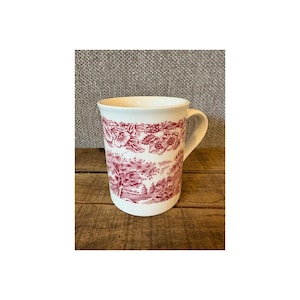 May include: A white ceramic mug with a red floral and landscape design. The mug has a curved handle and a cylindrical shape. The red design wraps around the mug, featuring trees, buildings, and floral patterns. The mug sits on a wooden surface.