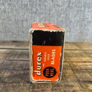 May include: A vintage box of Durex double edge blades. The box is orange with black accents and features the Durex logo and the words "double edge blades". The text "push here" is also visible on the box.