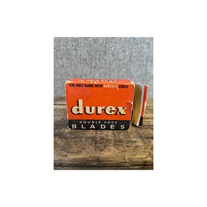 May include: An orange and black box of Durex Double Edge Blades. The box has the text "The only blade with Durexite edges" at the top. The box is sitting on a wooden surface.