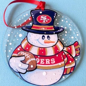 May include: A round, clear glass ornament featuring a painted snowman wearing a navy blue top hat with a red and gold 49ers logo. The snowman wears a red and gold striped scarf and holds a brown football. White dots simulate falling snow.