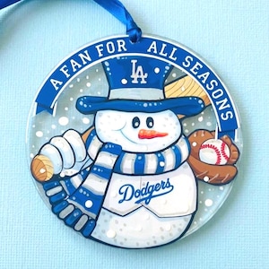 Könnte beinhalten: Ein rundes Glasornament mit einem Schneemann, der einen blauen Zylinder mit LA-Logo, einen blau-weiß gestreiften Schal und ein Dodgers-Trikot trägt. Der Schneemann hält einen Baseballschläger und einen Handschuh mit Baseball. Text: A FAN FOR ALL SEASONS.