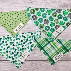 May include: Four green and white dog bandanas with different patterns. One bandana has a green shamrock pattern, one has a green plaid pattern, one has a green shamrock and donut pattern, and one has a green shamrock and pot of gold pattern. The bandanas are labeled with the names "Molly" and "Finley".