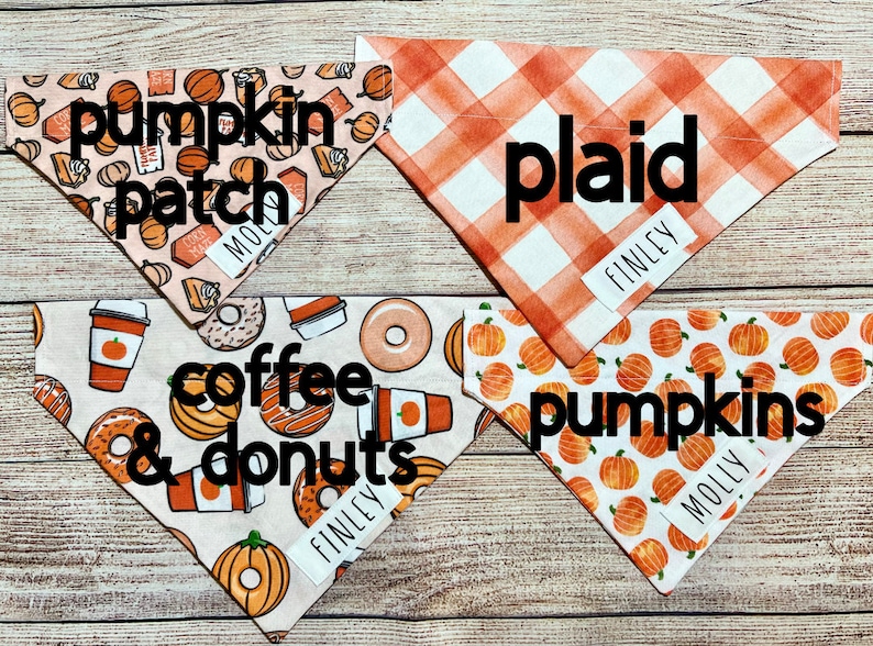 Op de afbeelding: Vier hondenbandana's met herfstthema's. Ontwerpen omvatten een pompoenveld, oranje ruit, koffie en donuts en pompoenen. Elke bandana heeft een wit label met de naam van een huisdier. De bandana's liggen op een houten oppervlak.