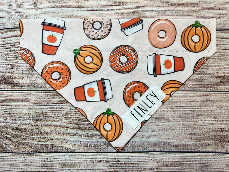 Op de afbeelding: Beige honden bandana met een patroon van donuts, pompoen kruidenlattes en pompoenen. De bandana heeft een wit label met "FINLEY" in zwart. Een accessoire voor huisdieren.