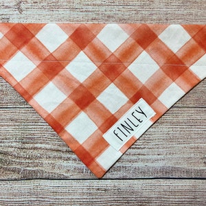 Op de afbeelding: Een driehoekige hondenbandana met een wit en oranje ruitpatroon. Een wit rechthoekig label met de naam "FINLEY" is op de bandana genaaid. De bandana is gemaakt van stof.