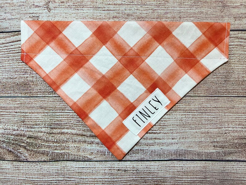 Op de afbeelding: Een driehoekige hondenbandana met een wit en oranje ruitpatroon. Een wit rechthoekig label met de naam "FINLEY" is op de bandana genaaid. De bandana is gemaakt van stof.