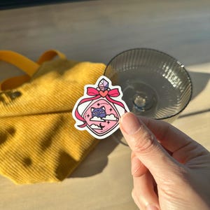 Può includere: Un adesivo con un design di bottiglia di pozione rosa con un gatto viola, stelle e nuvole. La bottiglia è adornata con un fiocco rosso e un cristallo. L'adesivo è tenuto vicino a una borsa di velluto a coste gialla e un bicchiere trasparente.