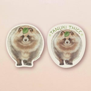 May include: Two stickers featuring a tanuki, a Japanese raccoon dog, with fluffy fur and a green leaf on its head. One sticker has the text "TANUKI THICCC" in a circular design. The stickers are on a light pink background.