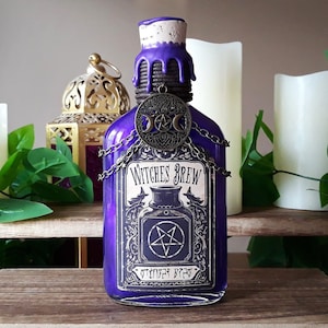 May include: A purple glass bottle with a black label that reads "Witches Brew". The bottle has a cork stopper and a metal chain with a pentagram charm. The bottle is sitting on a wooden surface.