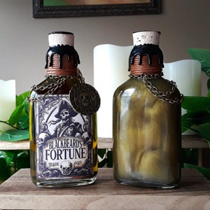 May include: Two glass bottles with gold liquid and black cork stoppers. The bottle on the left has a label that reads "Blackbeard's Fortune" with a pirate illustration. The bottle on the right has a gold chain around the neck.