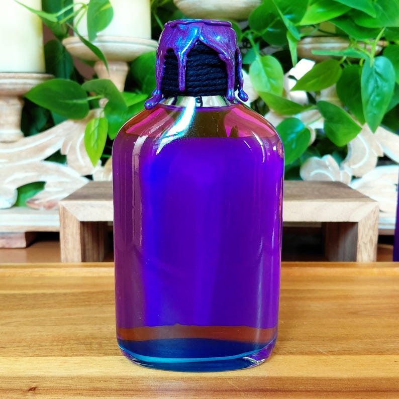 May include: A clear glass bottle filled with a vibrant purple liquid. The bottle has a decorative top with a dripped, iridescent purple finish. The bottle sits on a wooden surface, with green foliage in the background.