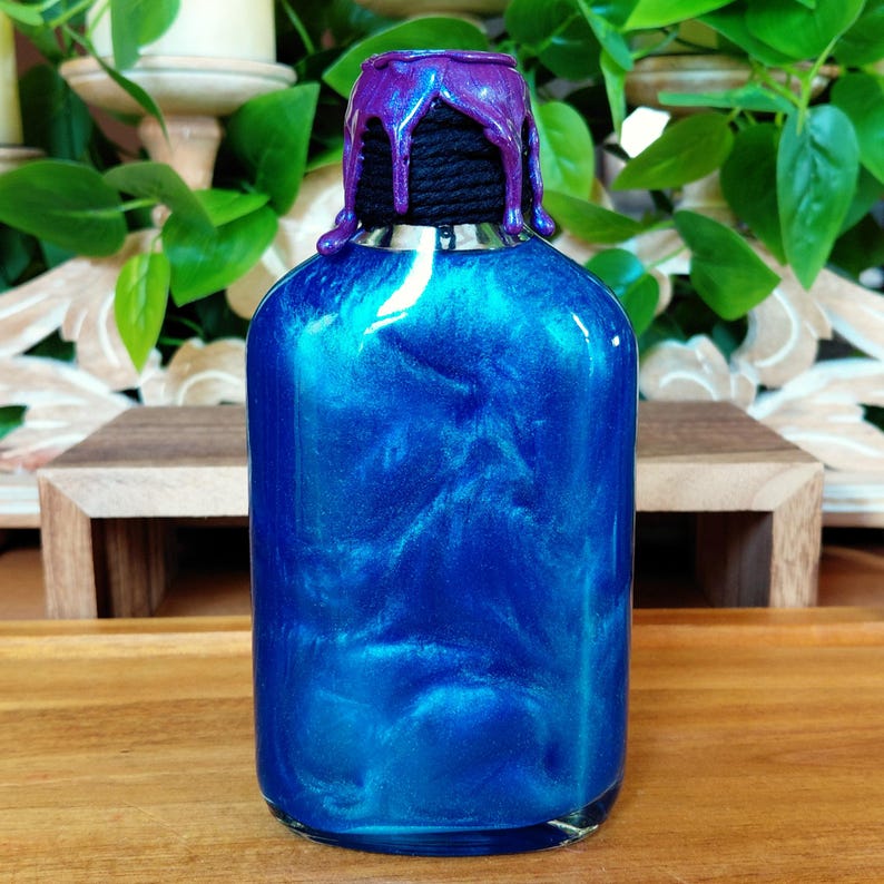 May include: A blue glass bottle with a shimmering, iridescent effect. The bottle has a black rope neck and a purple, dripped-wax-like top. The bottle is on a wooden surface, with green foliage in the background.