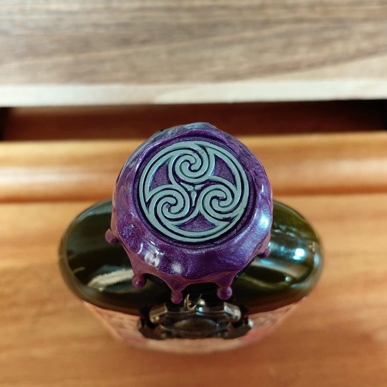 May include: A purple bottle stopper with a silver Celtic knot design. The stopper has a metallic sheen and is dripping down the sides. The bottle is green and has a decorative metal clasp.