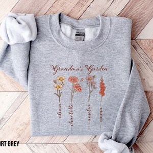 May include: A gray sweatshirt with the text "Grandma's Garden" and four flowers with names written below them: Olivia, Charlotte, Amelia, Emma.
