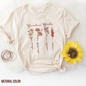 May include: A beige t-shirt with the text "Grandma's Garden" printed on the front. The text is in a brown font. There are four stylized floral designs with names written underneath each flower: Olivia, Charlotte, Amelia, and Emma.
