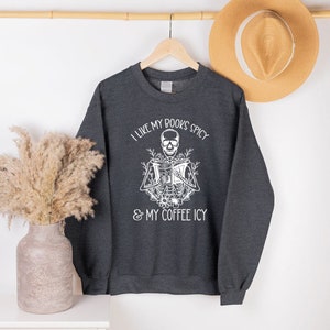 May include: Dark gray sweatshirt with the text "I LIKE MY BOOKS SPICY & MY COFFEE ICY" and a white skeleton graphic reading a book. The sweatshirt is hanging on a wooden hanger with a straw hat in the background.