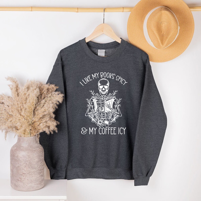 May include: Dark gray sweatshirt with the text "I LIKE MY BOOKS SPICY & MY COFFEE ICY" and a white skeleton graphic reading a book. The sweatshirt is hanging on a wooden hanger with a straw hat in the background.