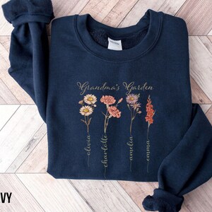 May include: Navy blue sweatshirt with a floral design and the text "Grandma's Garden". The names Olivia, Charlotte, Amelia, and Emma are written vertically below the flowers.