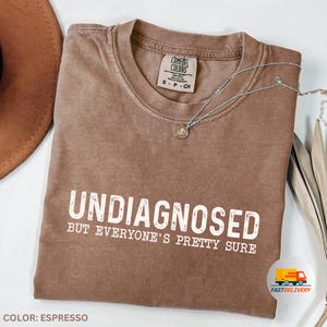 Könnte beinhalten: Espresso-braunes T-Shirt mit dem Text "Undiagnosed but everyone's pretty sure" in Weiß. Das Shirt ist aus einem weichen, bequemen Material.