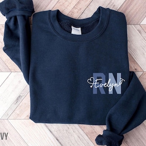 Pode incluir: Camisola azul marinho com um gráfico branco que diz "RN Evelyn" com um estetoscópio em forma de coração.