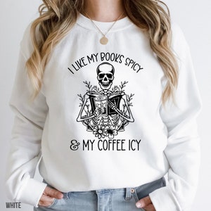 May include: White sweatshirt with a black graphic of a skeleton reading a book and holding a drink. The text reads, "I LIKE MY BOOKS SPICY & MY COFFEE ICY."