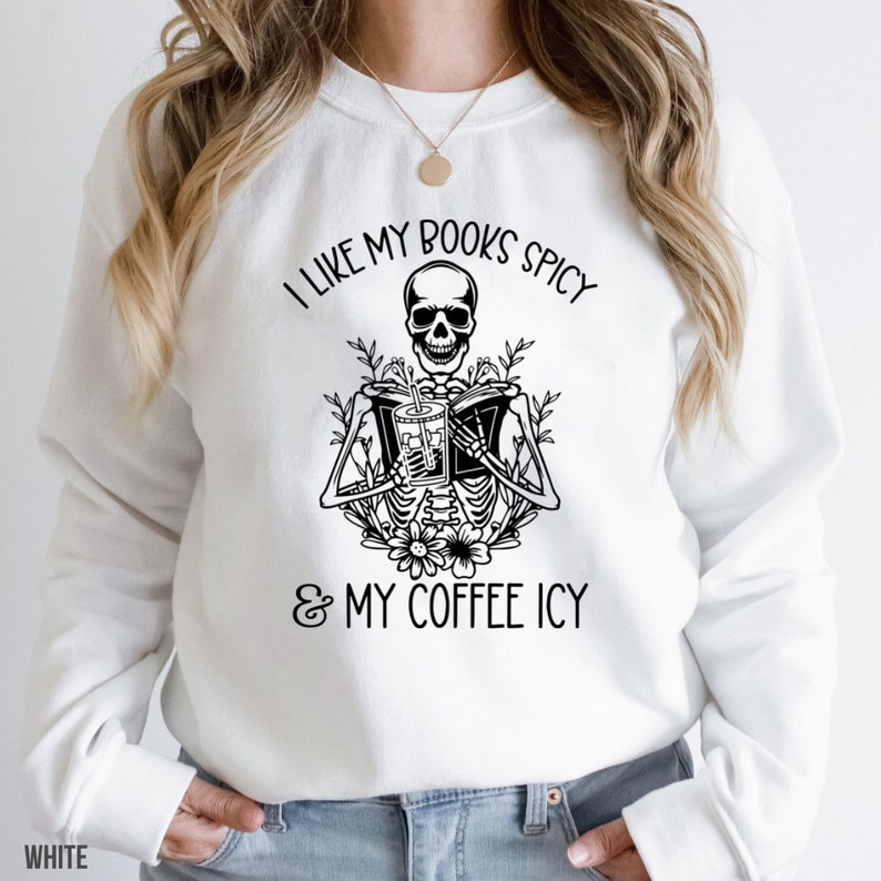 May include: White sweatshirt with a black graphic of a skeleton reading a book and holding a drink. The text reads, "I LIKE MY BOOKS SPICY & MY COFFEE ICY."