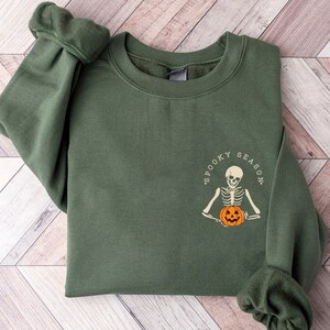 May include: A dark green sweatshirt with a white graphic of a skeleton holding a jack-o'-lantern. The graphic says "Spooky Season".