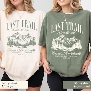 May include: Two t-shirts, one cream and one khaki green, featuring a mountain and forest design. The text on the shirts reads "LAST TRAIL before the veil" and "Emma's Bachelorette, Gatlinburg, Tennessee, EST. 2025."