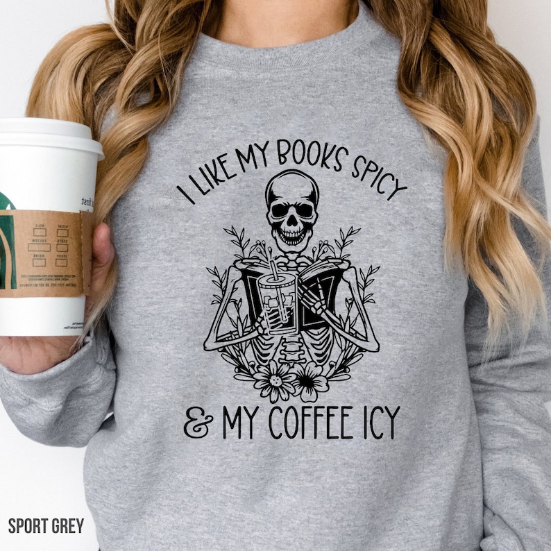 May include: Sport grey sweatshirt with a black graphic of a skeleton reading a book and holding a drink. The text reads "I LIKE MY BOOKS SPICY & MY COFFEE ICY". The sweatshirt is a crew neck style.
