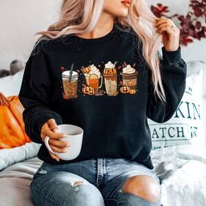 May include: A black sweatshirt with a graphic of four fall-themed coffee drinks with pumpkin spice, whipped cream, and fall leaves. The sweatshirt is being worn by a person sitting on a couch.