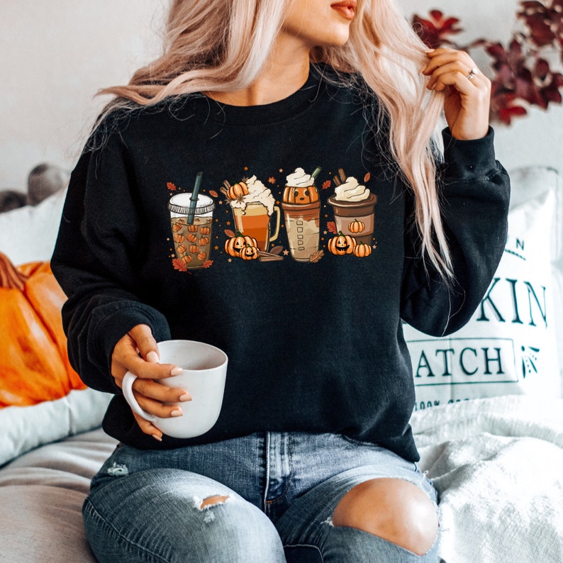 May include: A black sweatshirt with a graphic of four fall-themed coffee drinks with pumpkin spice, whipped cream, and fall leaves. The sweatshirt is being worn by a person sitting on a couch.