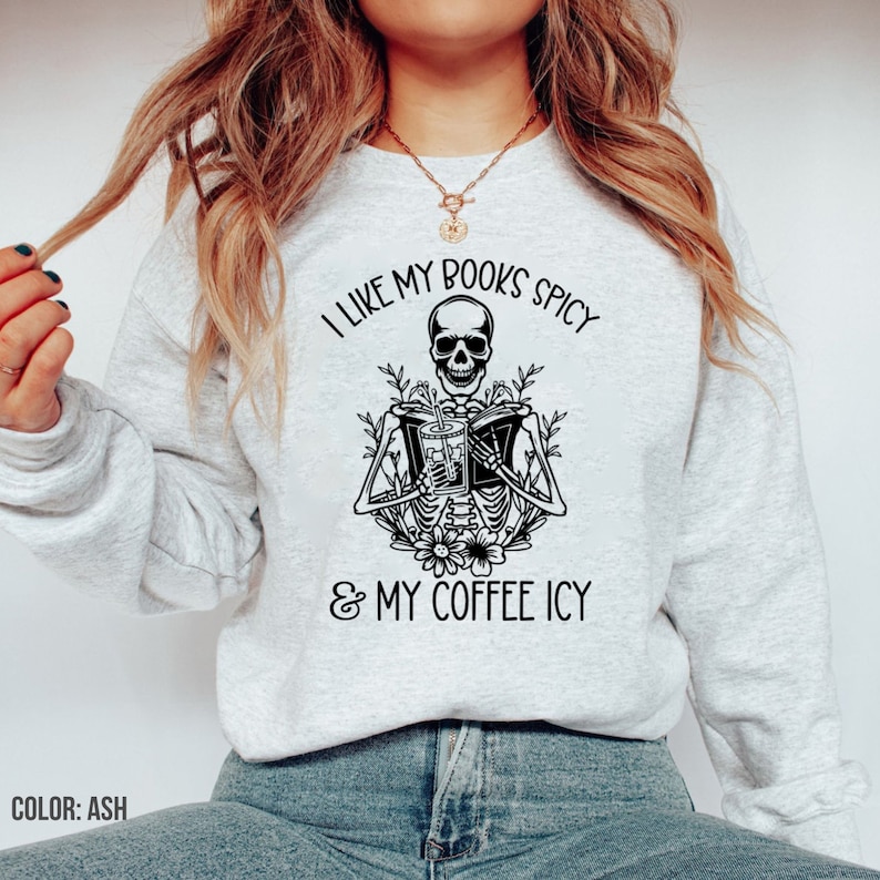 May include: Ash-colored sweatshirt featuring a black graphic of a skeleton reading a book and holding a drink. The text reads, "I LIKE MY BOOKS SPICY & MY COFFEE ICY." The sweatshirt is a casual, comfortable style.