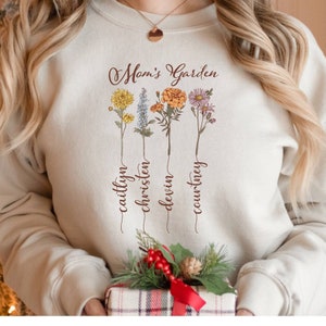 May include: A beige sweatshirt with the text "Mom's Garden" and four floral designs with the names "Caitlyn", "Christen", "Devin", and "Courtney" written underneath each flower.