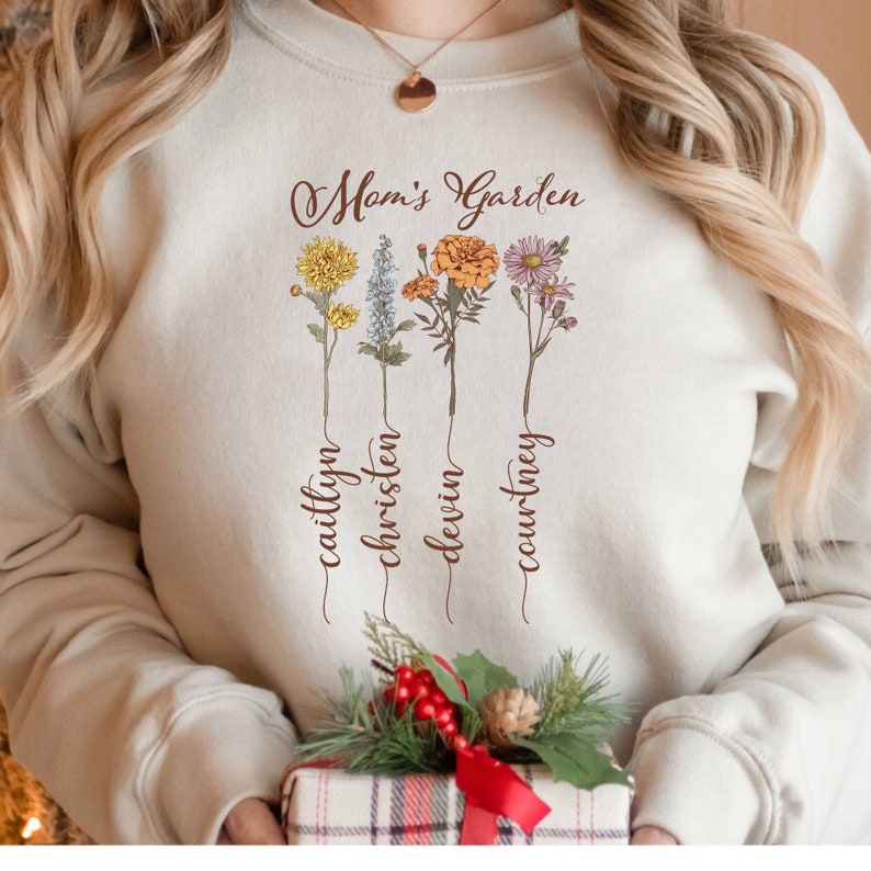 May include: A beige sweatshirt with the text "Mom's Garden" and four floral designs with the names "Caitlyn", "Christen", "Devin", and "Courtney" written underneath each flower.
