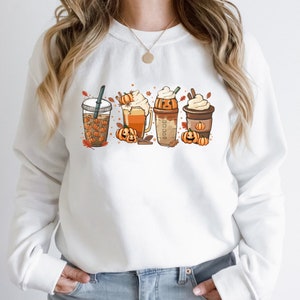 May include: White sweatshirt with a graphic of four fall-themed drinks with pumpkin faces. The drinks include a pumpkin spice latte, a pumpkin beer, a pumpkin milkshake, and a pumpkin iced coffee.